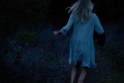 Woman in dress runs down a dark wooded path.