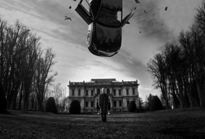 A car hanging in the sky above a man standing with a mansion behind him